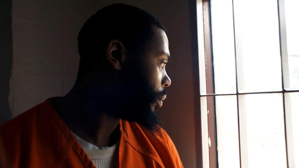 A man in prison clothing looks out a barred window.