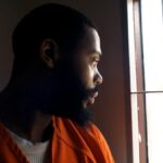 A man in prison clothing looks out a barred window.