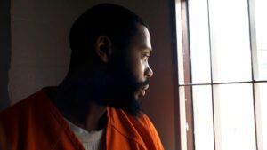 A man in prison clothing looks out a barred window.