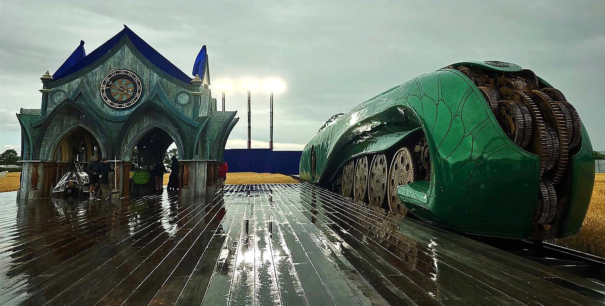 A rounded green train sits on the track near a unique-looking station.