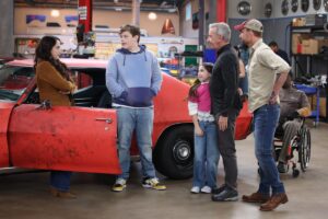 A woman standing in the doorway of a dirty red car as two children and two men look in her direction.