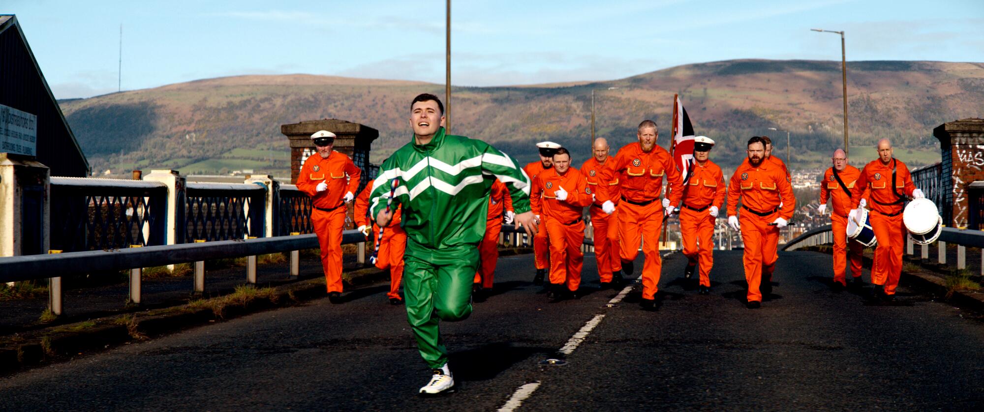 A man in a green track suit runs down a street followed by others in orange in "Kneecap."