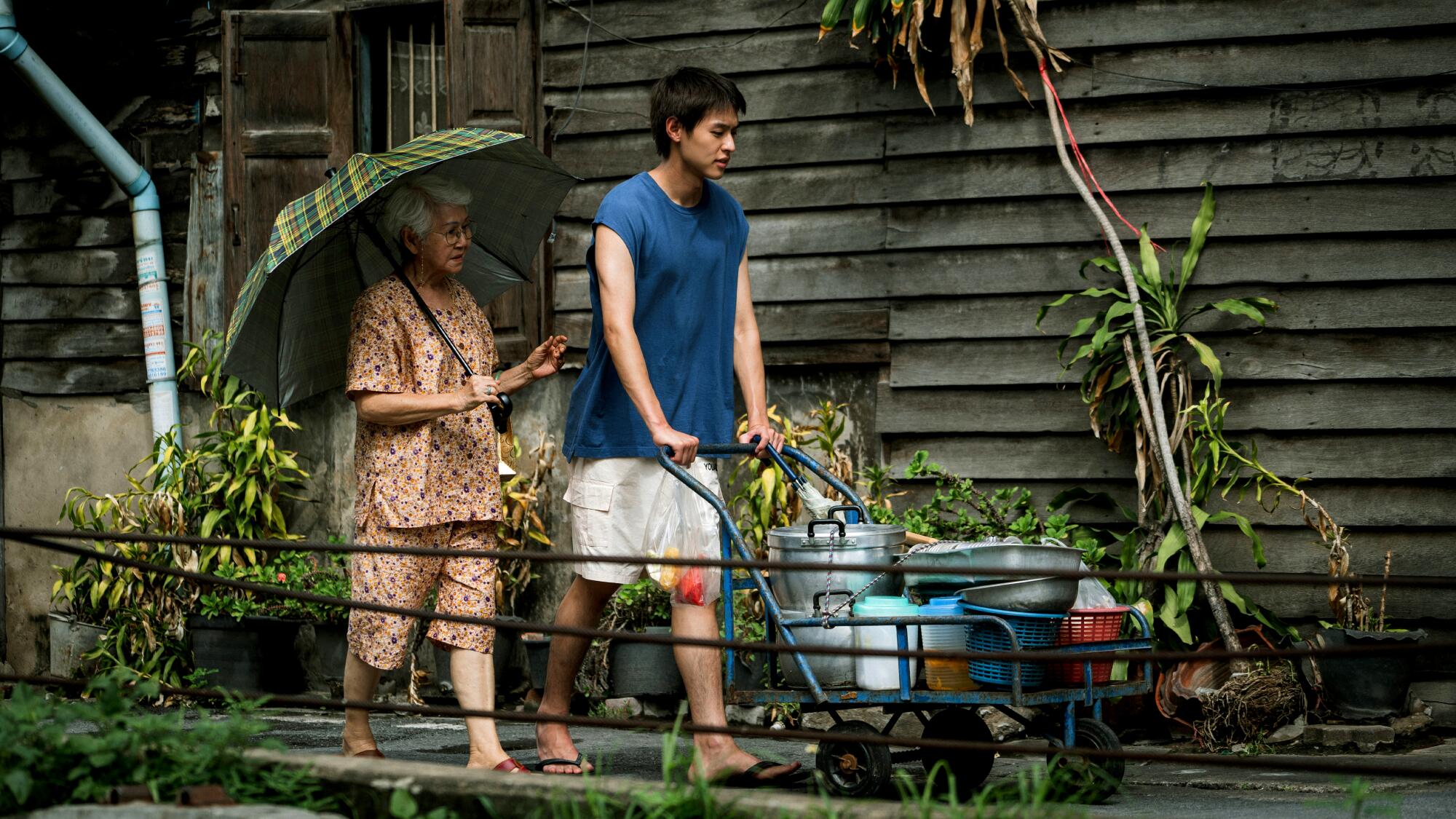 A young man pushes a cart filled with pots while his grandmother walks beside him under an umbrella in the film. 