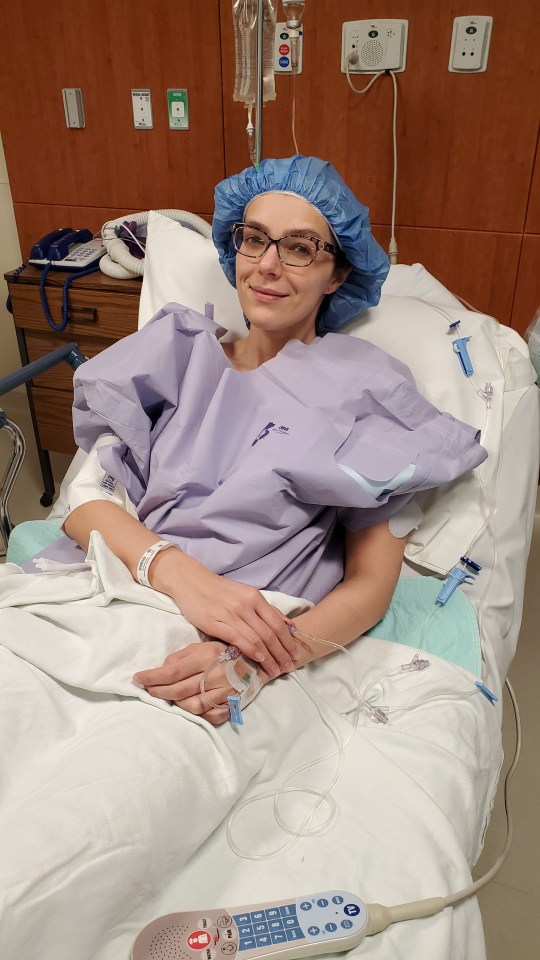 Adrienne Curry in a hospital gown and surgical cap in a hospital bed.