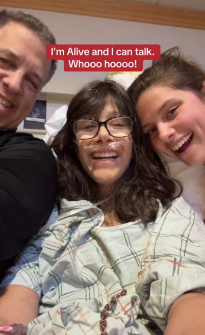 A woman in a hospital bed with her family, smiling and saying "I'm alive and I can talk."