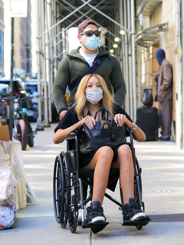 Wendy Williams in a wheelchair, wearing a mask, being pushed down a city street.