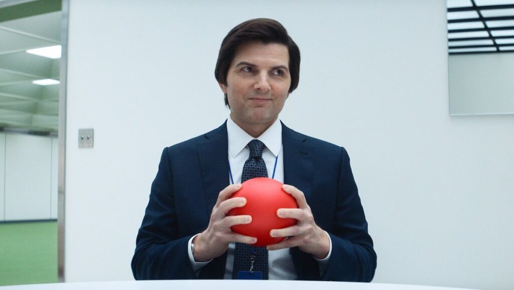 A smirking Mark holds a red ball sitting at a table on Severance