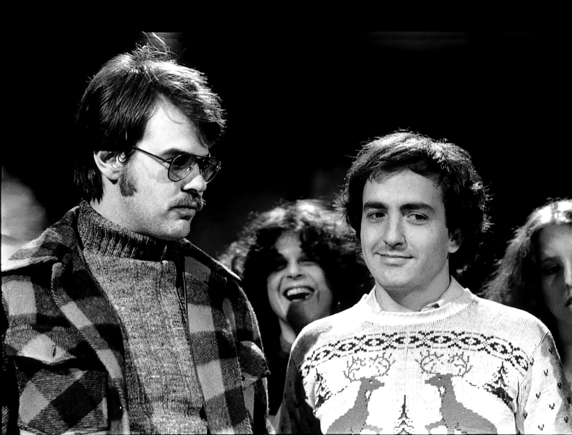 Two men in sweaters standing next to each other. The man on the right is smiling.