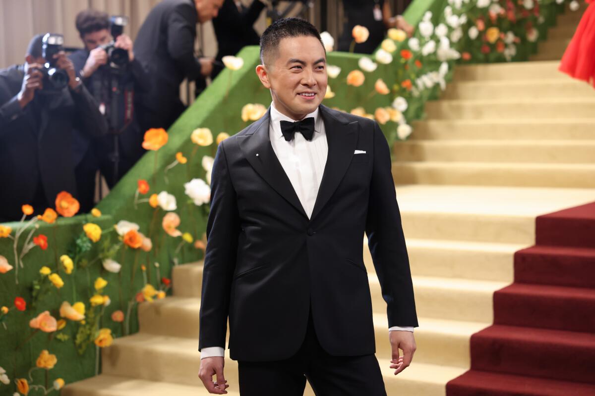 A man in a black tuxedo stand near a set of stairs covered in cream and red carpeting.