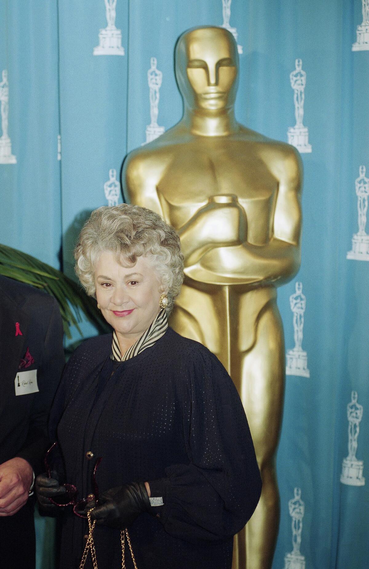 Joan Plowright in a black coat standing in front of a large, golden Oscars statue