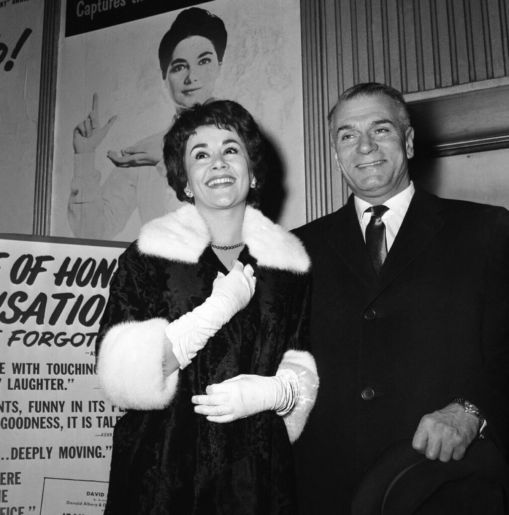 Black and white photo of actor Joan Plowright in a coat and long white gloves standing next to actor Laurence Olivier