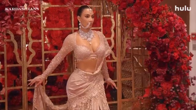 Kim Kardashian in a beaded, light-pink sari in front of a red floral display.