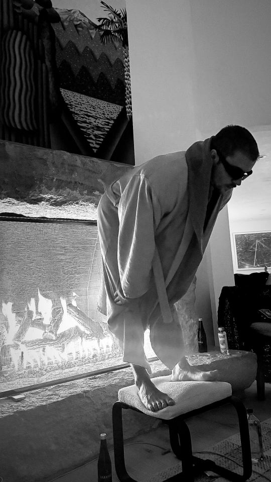 Black and white photo of a man in a robe balancing on a small stool in front of a fireplace.