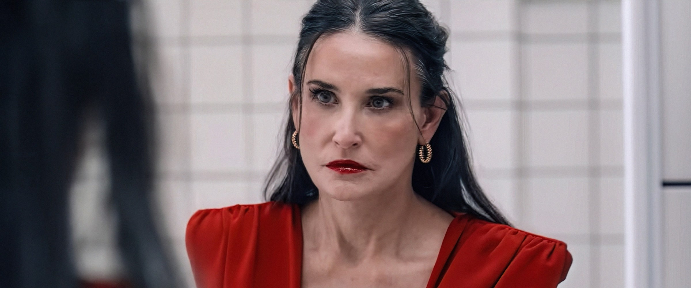 Demi Moore, wearing a red dress, looks in the mirror in The Substance