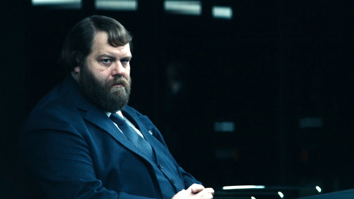 The intimidating Mr. Drummond sits at a board table in a suit with his arms crossed on Severance