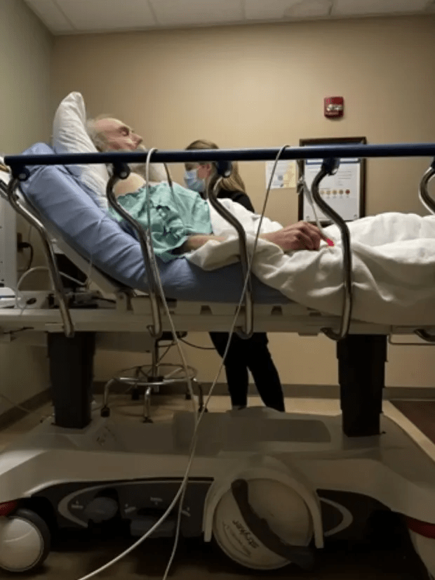 Elderly man lying in a hospital bed, attended by a healthcare worker.