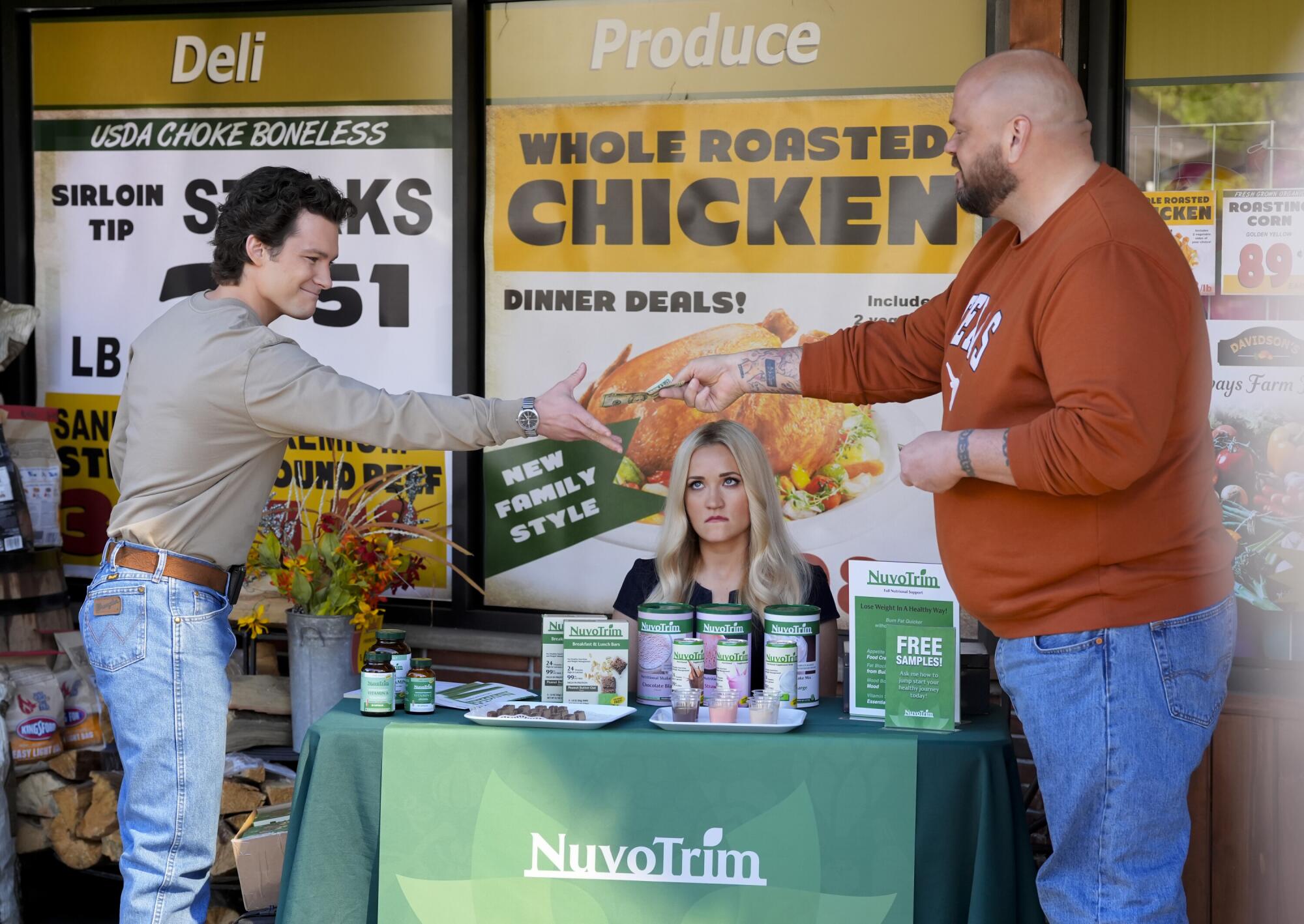 Two men standing exchange money over a woman sitting at table covered in a green cloth and sign that reads "NuvoTrim."