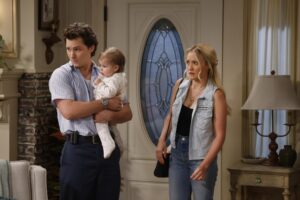 A man holding a baby stands next to a woman in a denim vest and jeans near a doorway.