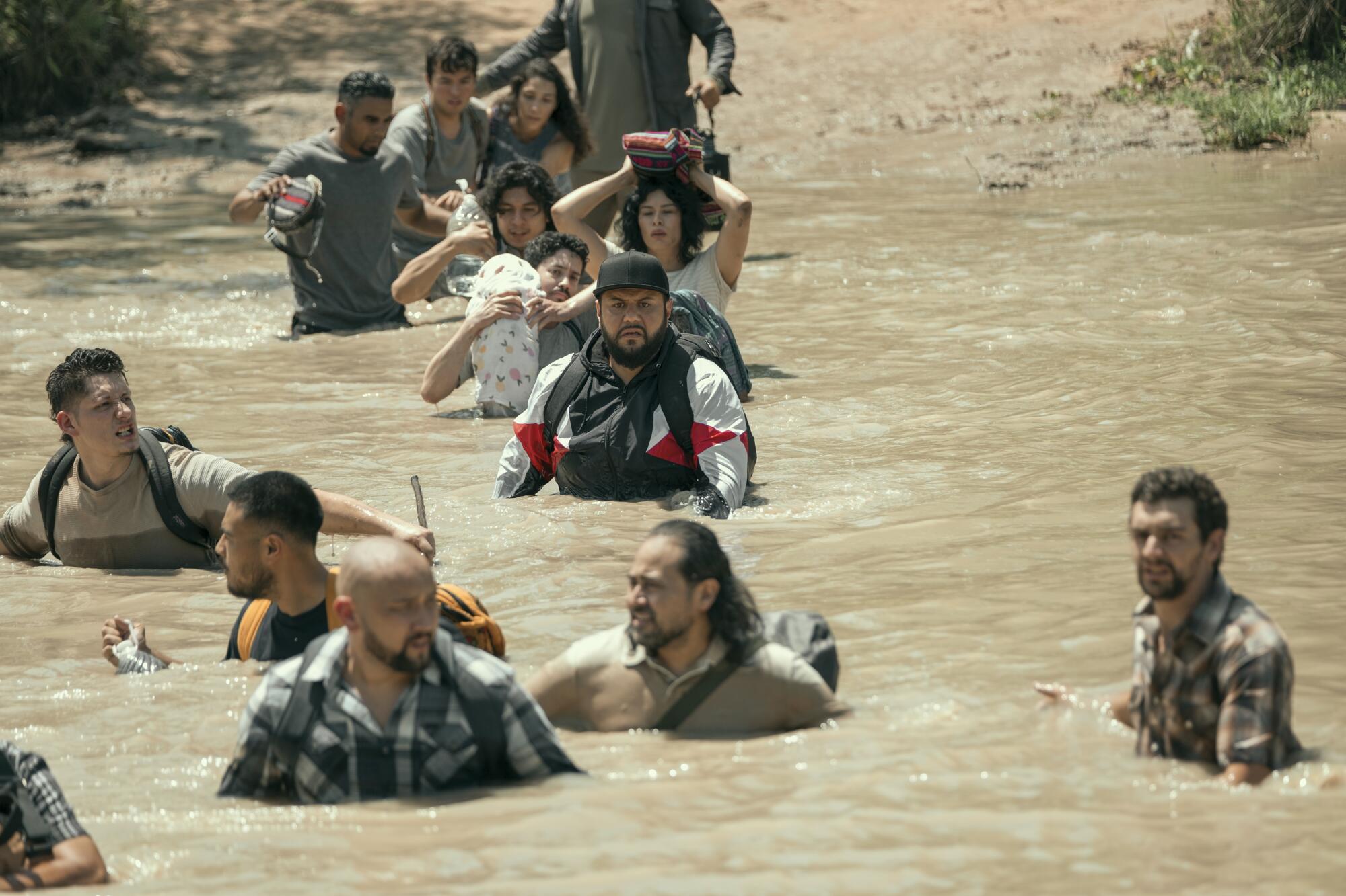 Men and women wade through a brown river at a border crossing. 