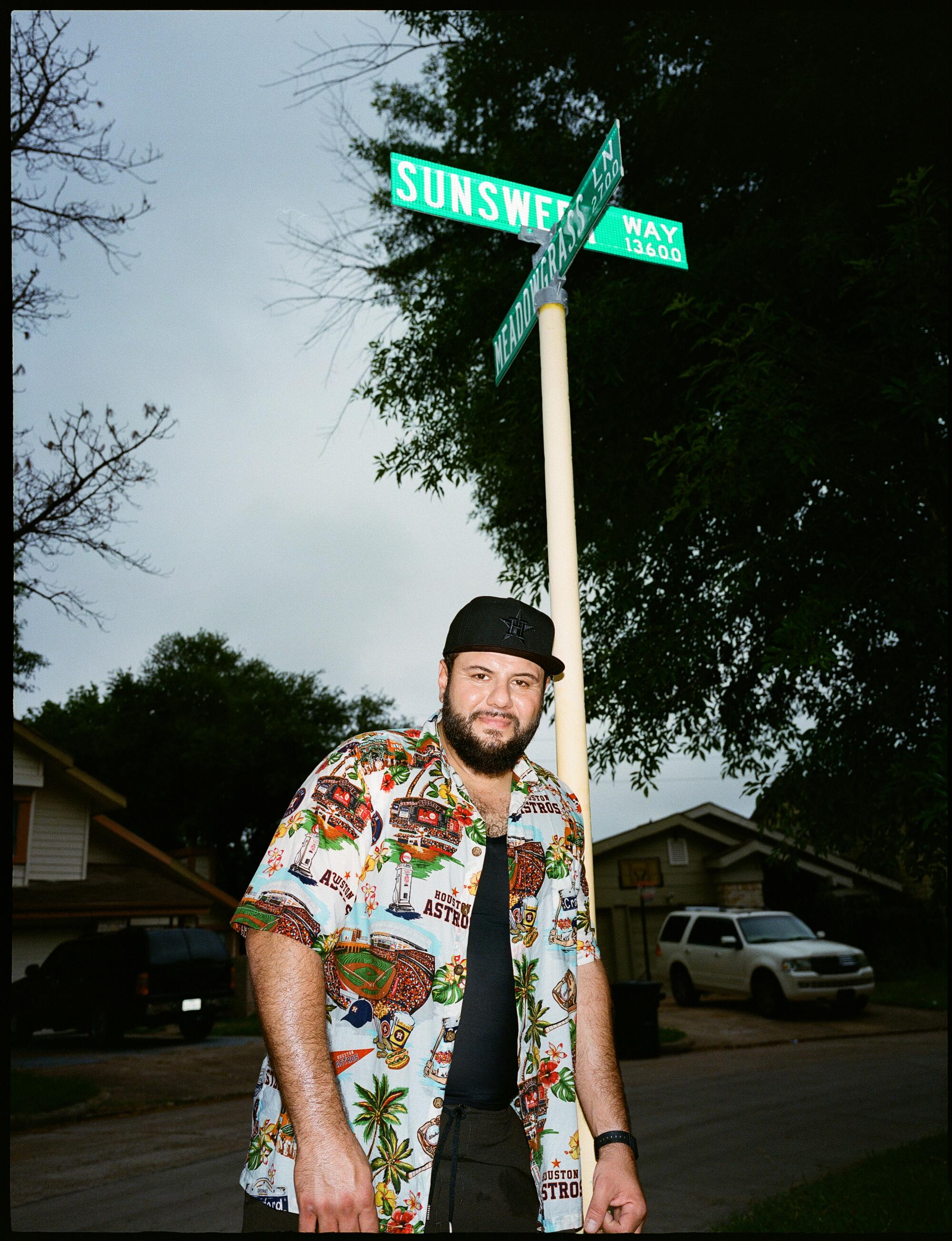 A man in a black ball cap and floral shirt leans against a street sign post.