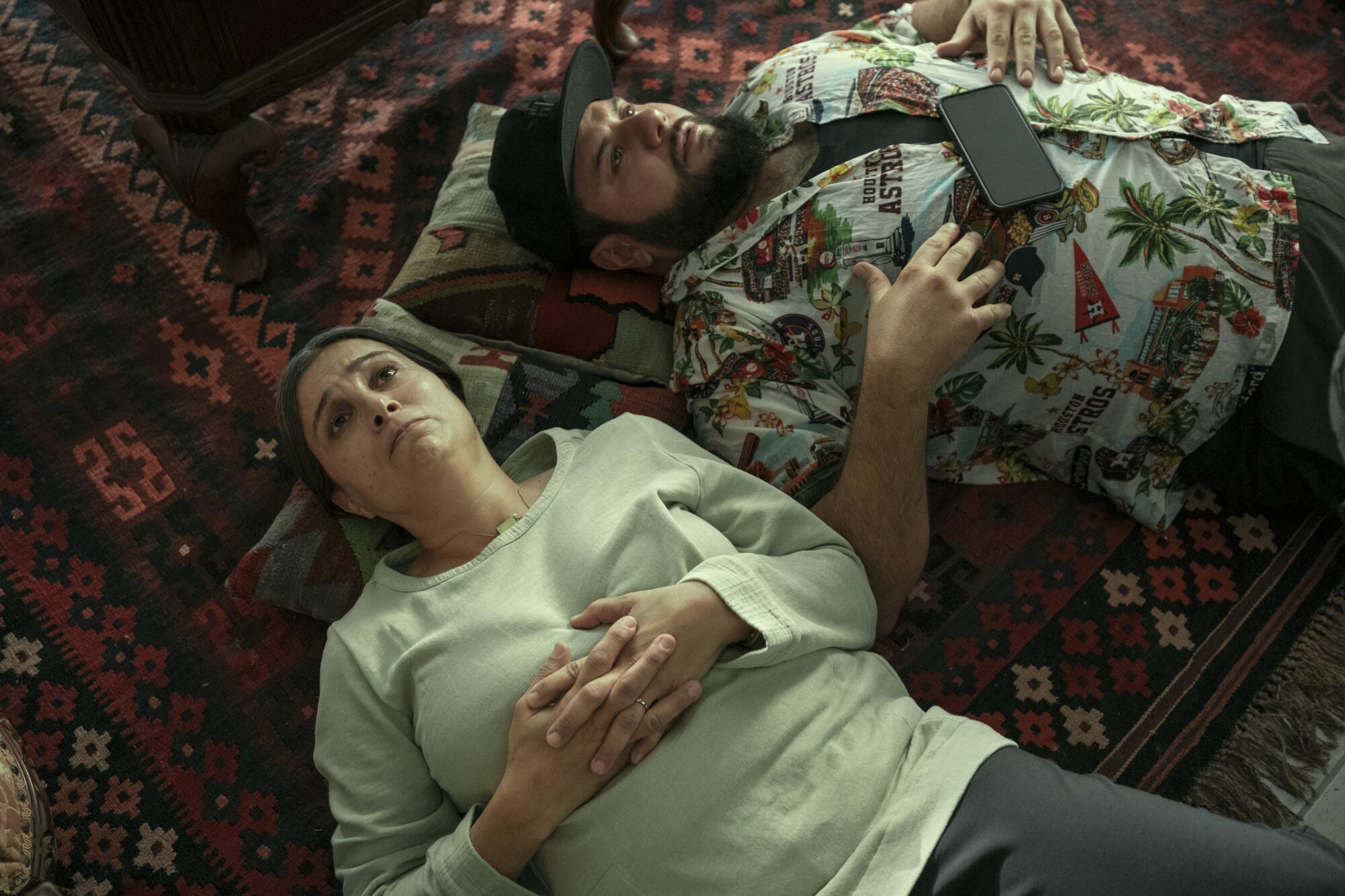 A woman in white shirt and gray pants lies face up on a rug next to a man with a beard in black ball cap and floral shirt.