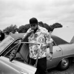 A man in a ballcap and floral shirt sips from a tea cup in the doorway of a car with its trunk open.