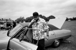 A man in a ballcap and floral shirt sips from a tea cup in the doorway of a car with its trunk open.