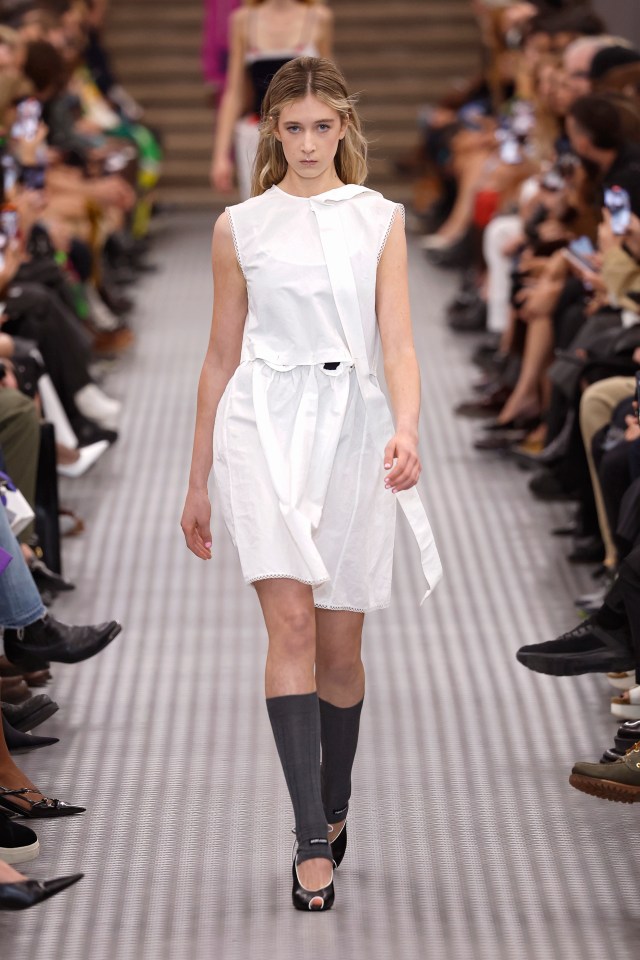 Model walking a runway in a white dress and dark socks.