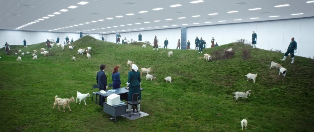 People near a small desk looking out at an interior pasture of green grass, goats, and herders on Severance