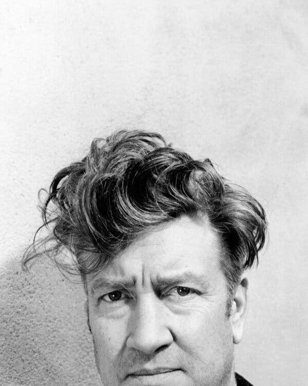 A black and white photograph of a young David Lynch standing against a wall. He is positioned toward the bottom of the frame and his chin is cut off slightly.