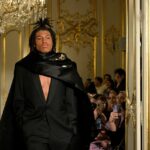 Model in black suit and cape walking a runway.