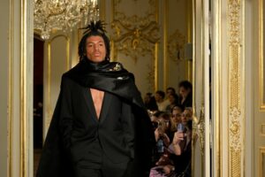 Model in black suit and cape walking a runway.