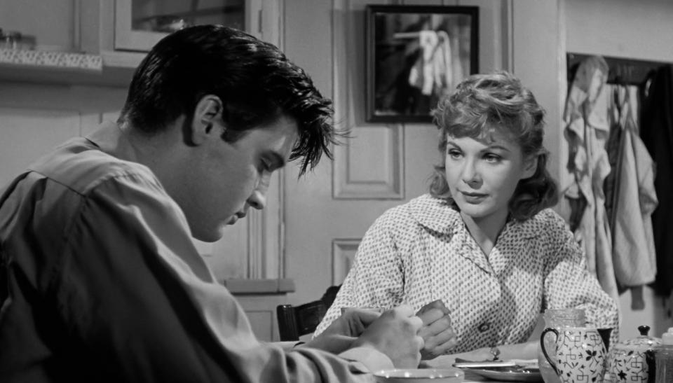 Black and white still image from the film *King Creole* showing Elvis Presley and Jan Shepard seated at a table.