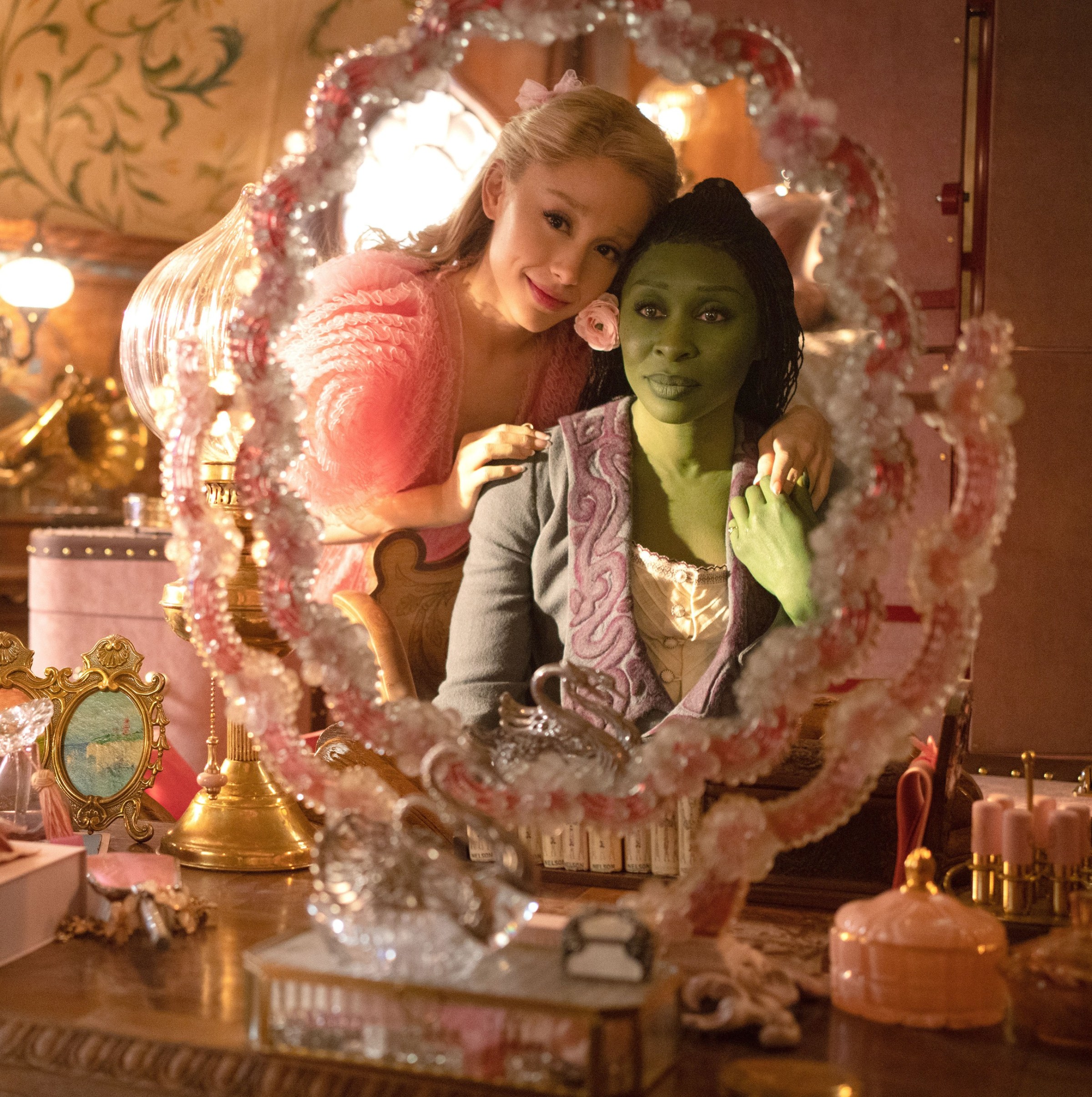 Glinda (Ariana Grande) presses her head against Elphaba’s (Cynthia Erivo) as they look into a mirror lined with pink frills in Wicked: Part I