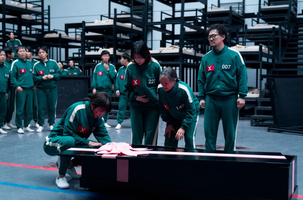 A group of remaining Squid Game contestants gather around a gift-wrapped coffin in a still from season 3