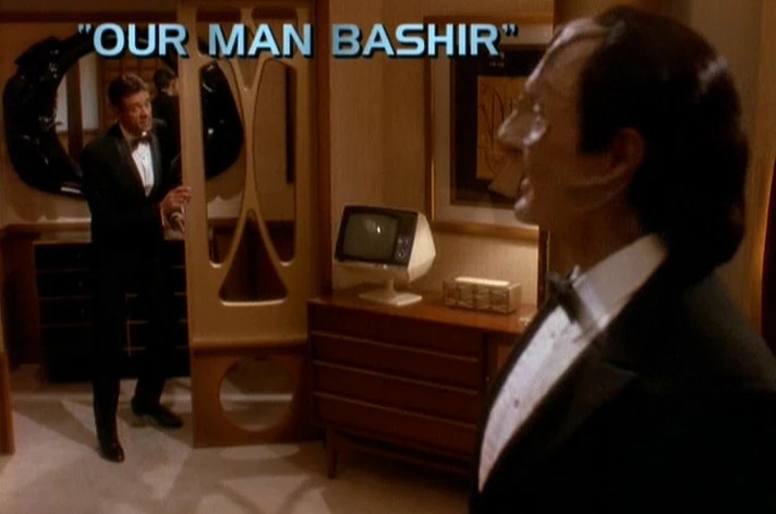 Julian Bashir and Elim Garak stand around in a cool mid-century hotel room (on the holodeck) wearing tuxedos. The title card “Our Man Bashir” is visible.