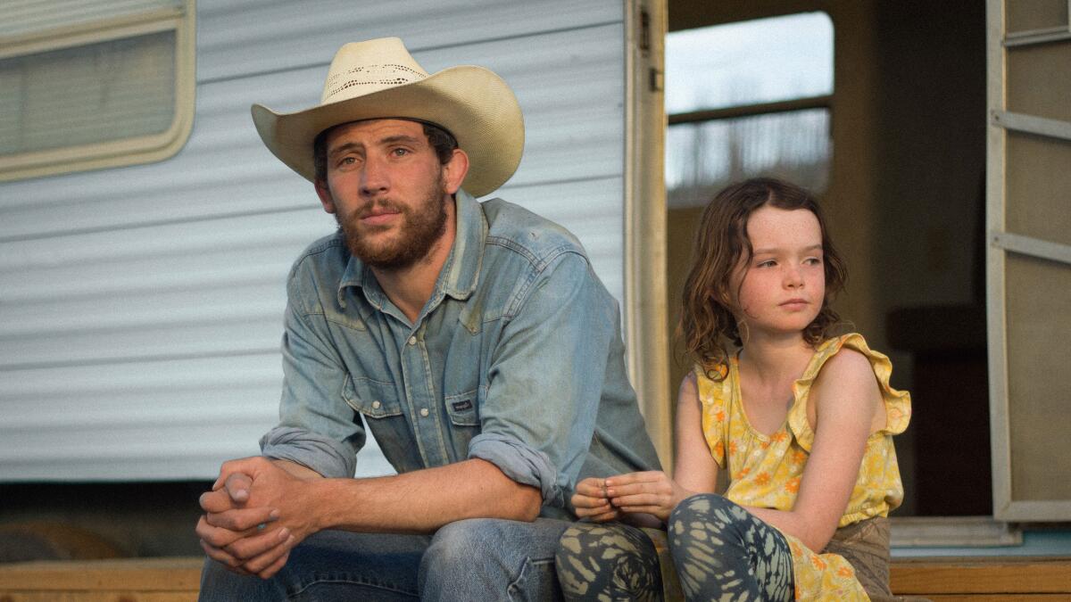 A man in a cowboy hat and his daughter sit outside the door of their trailer.