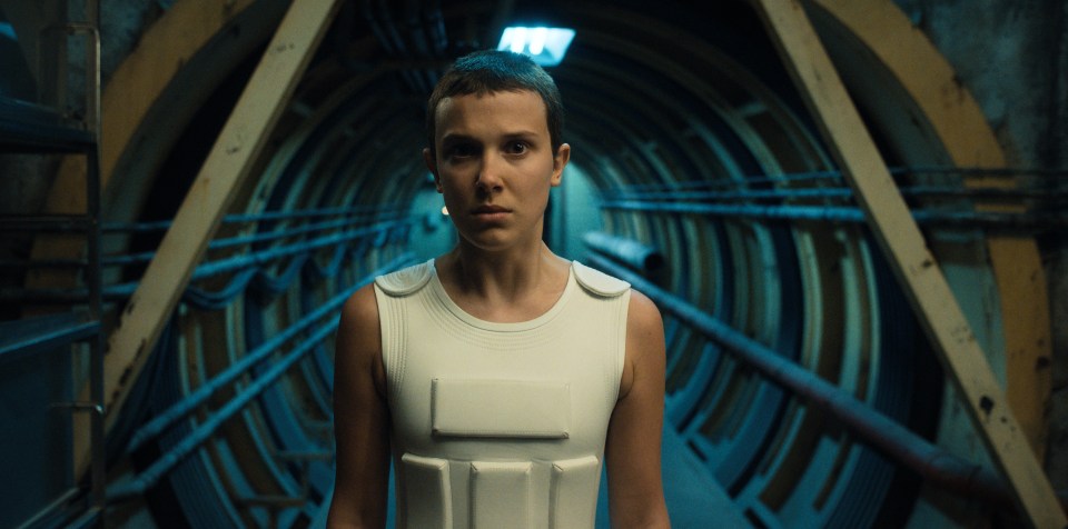 Millie Bobby Brown as Eleven in Stranger Things, wearing a white protective vest in a tunnel.