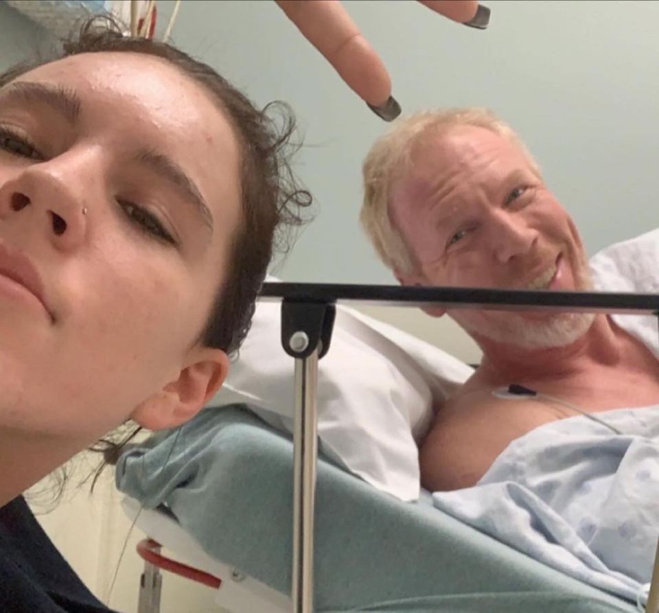 A man in a hospital bed smiles at the camera while his daughter playfully makes a hand gesture above his head.