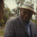 A man in a straw hat and plaid blazer smiles as he looks at a woman.