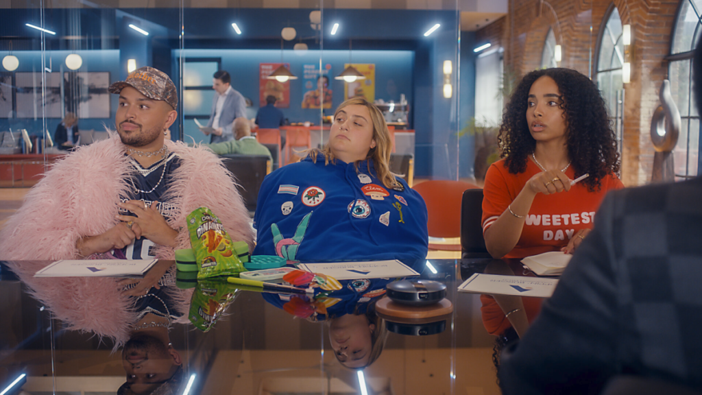 A man in a pink feathery coat, a woman in a blue hoodie and a woman in red shirt sit at a conference table.