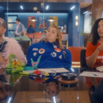 A man in a pink feathery coat, a woman in a blue hoodie and a woman in red shirt sit at a conference table.