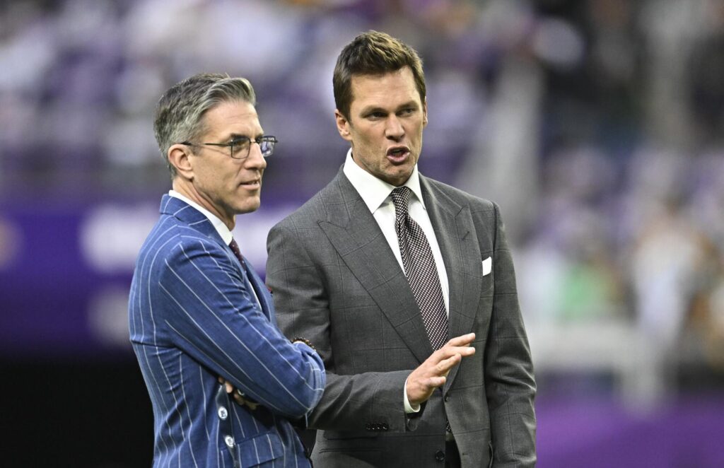 Kevin Burkhardt and Tom Brady stand next to each other on a field.