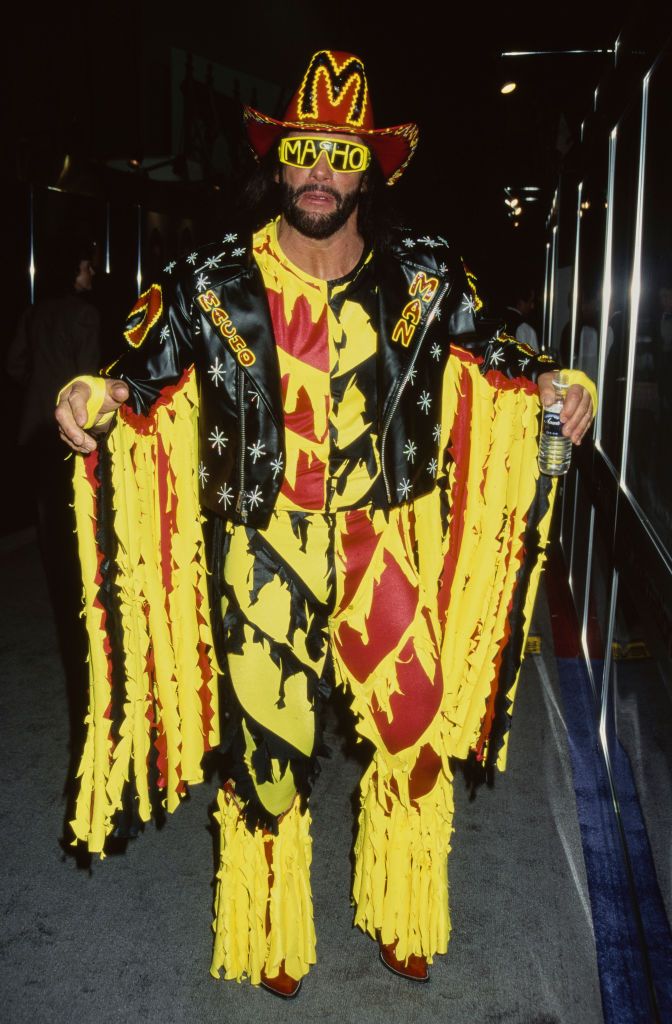 Macho Man Randy Savage wearing a black, yellow, and red fringed outfit with a matching cowboy hat and sunglasses, walking at an event.