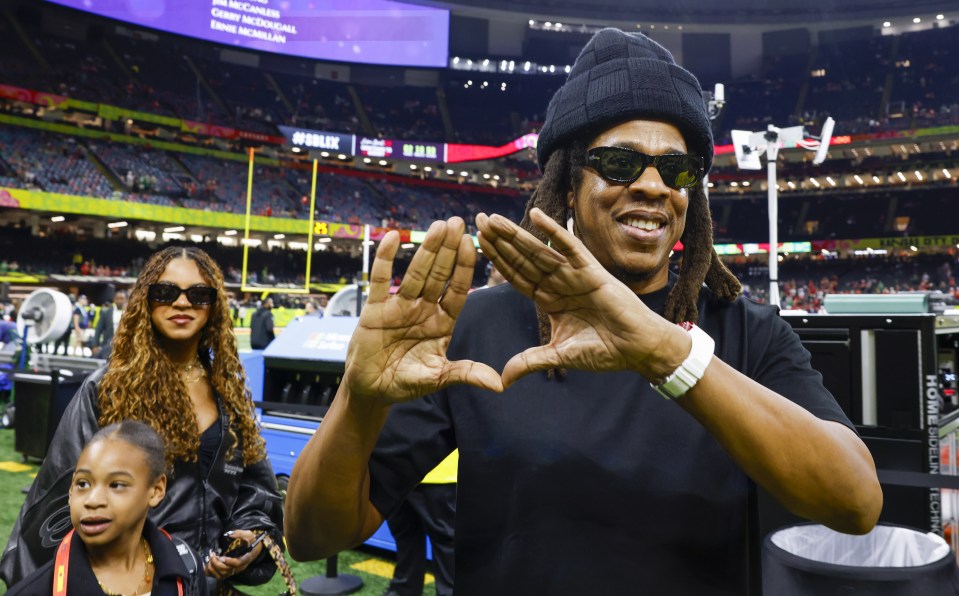 Jay-Z making a heart shape with his hands at a Super Bowl game with his daughters.