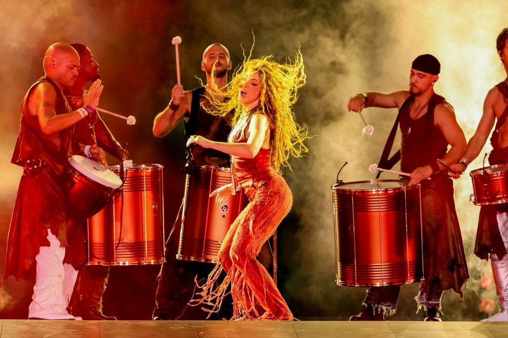 Shakira dances on stage in a red outfit, surrounded by drummers playing large drums, with golden lighting enhancing the high-energy atmosphere.