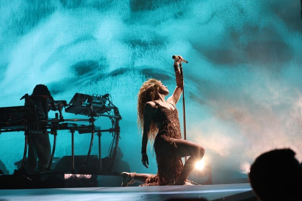 Shakira kneels on stage, singing into a microphone with a powerful pose, as blue lighting and a dramatic backdrop set the mood.