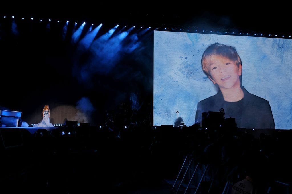 Shakira stands on stage near a piano, performing in front of a giant screen displaying a photo of her son, surrounded by blue lighting.