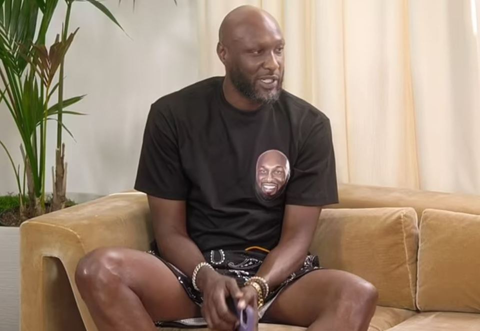 Lamar Odom sitting on a couch, wearing a black t-shirt with his own face on it.