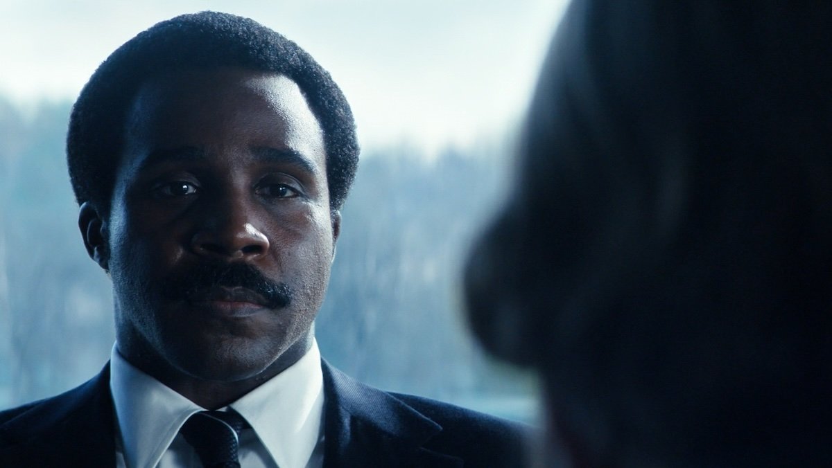 Seth Milchick in a suit sitting at a table with a snowy background behind the windows on Severance